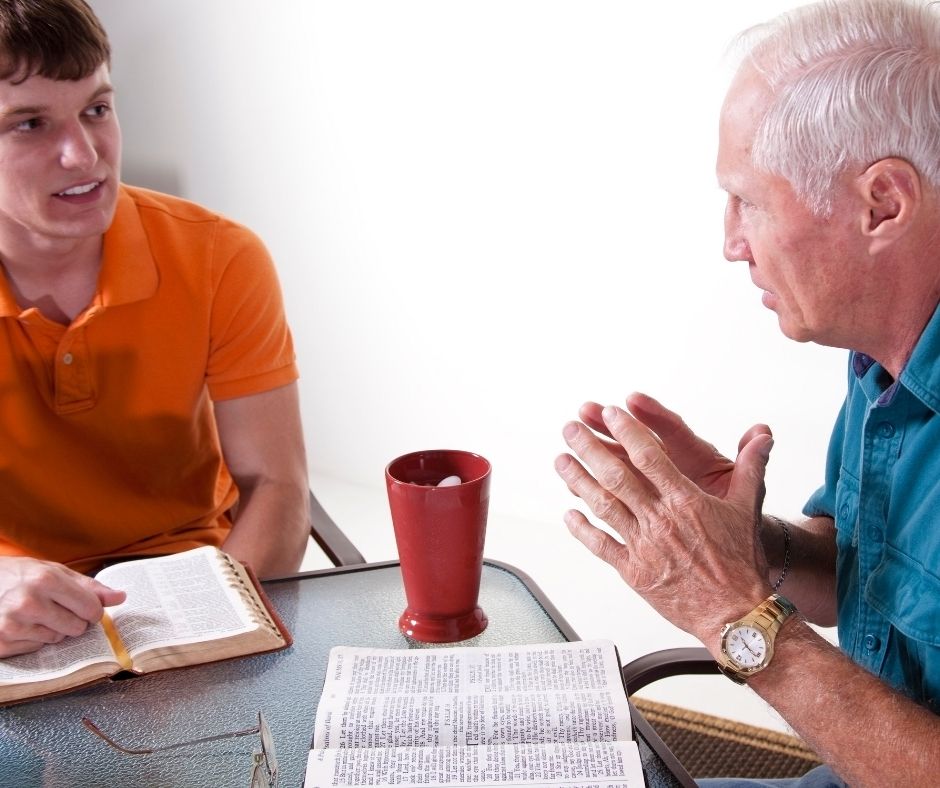 young man and senior man sitting a table with open Bible discussing scripture